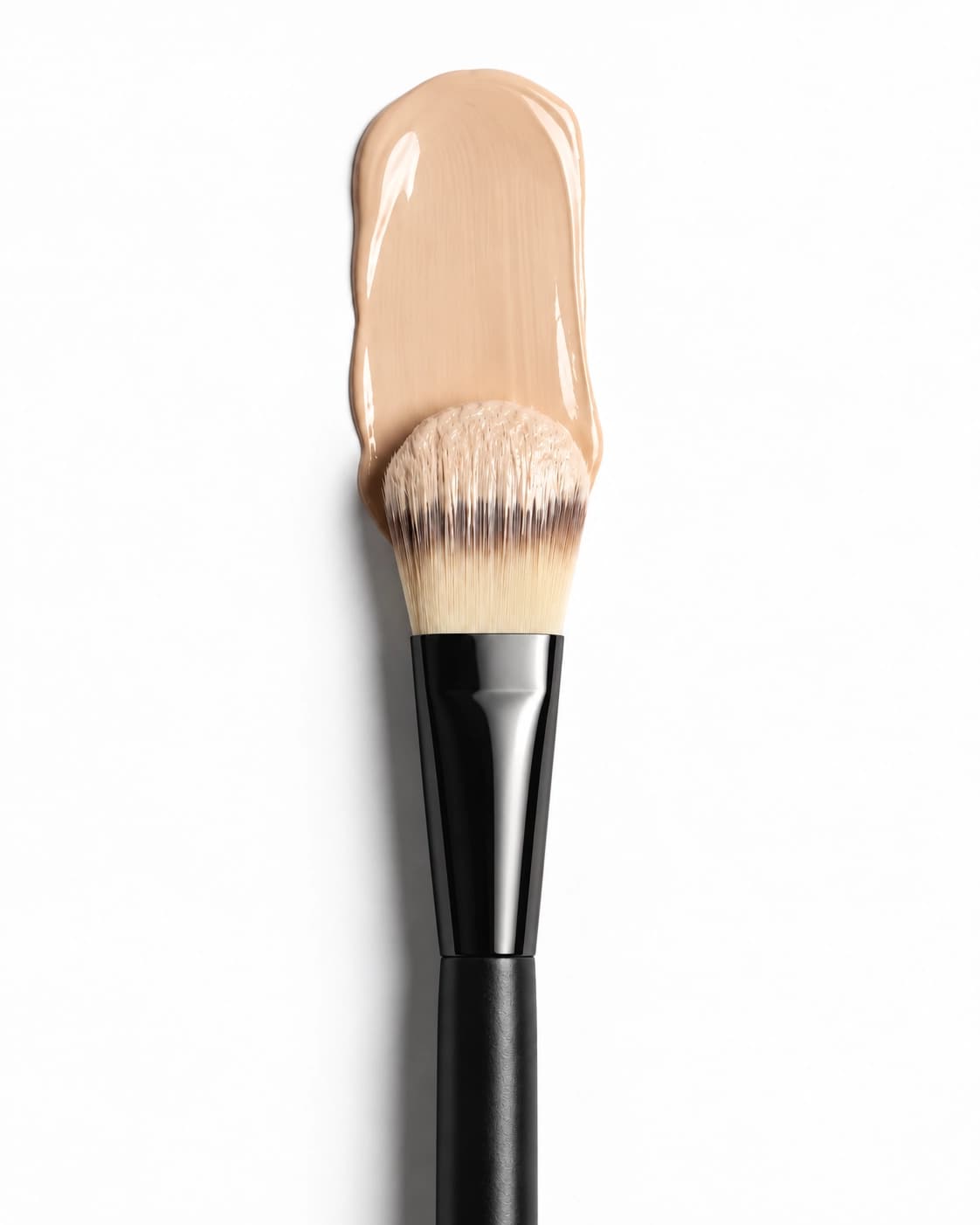 Brocha para base de maquillaje junto a una muestra de base líquida sobre un fondo de estudio
