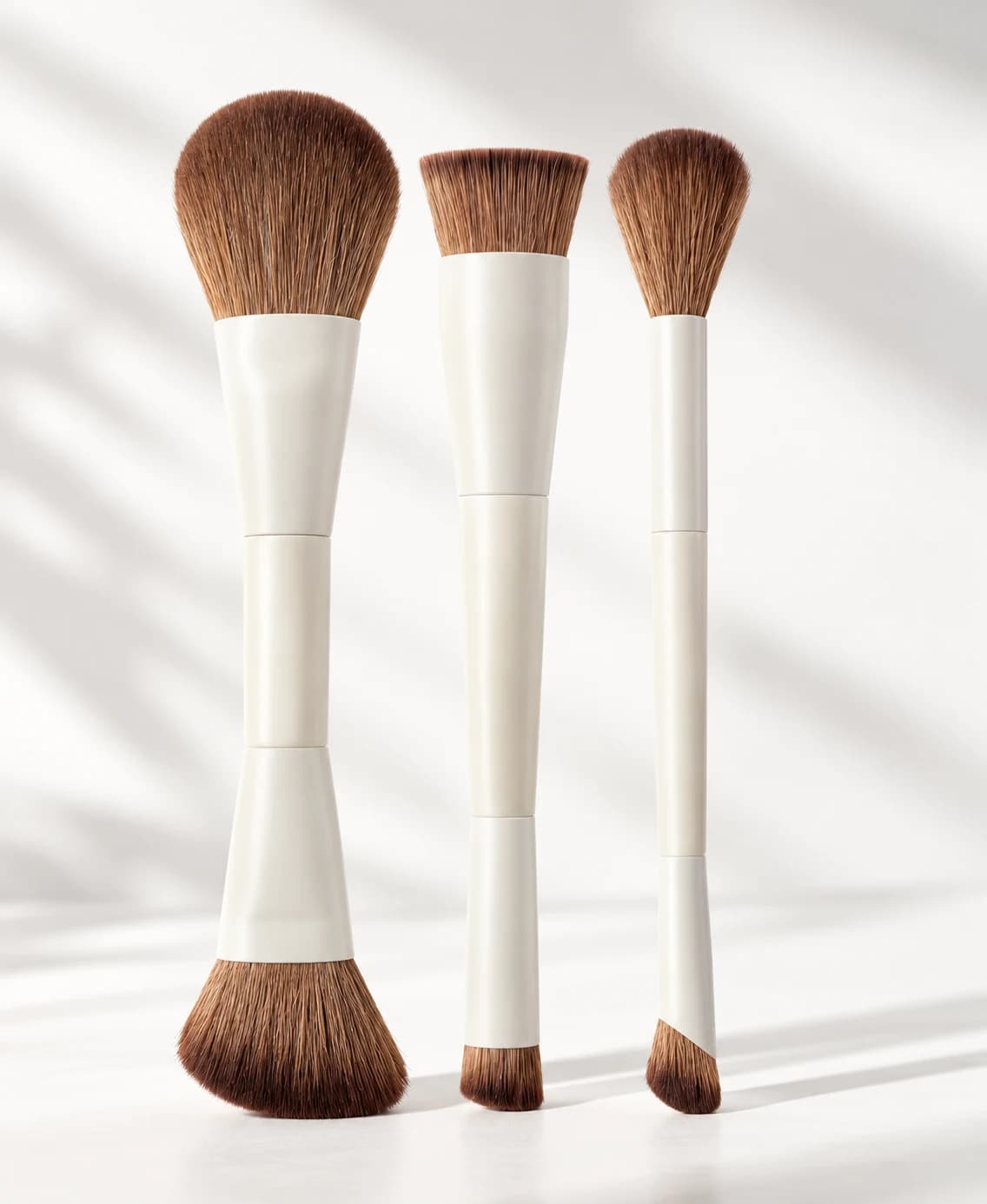 Three ivory-colored double-ended makeup brushes with brown bristles standing upright, showing six brush heads