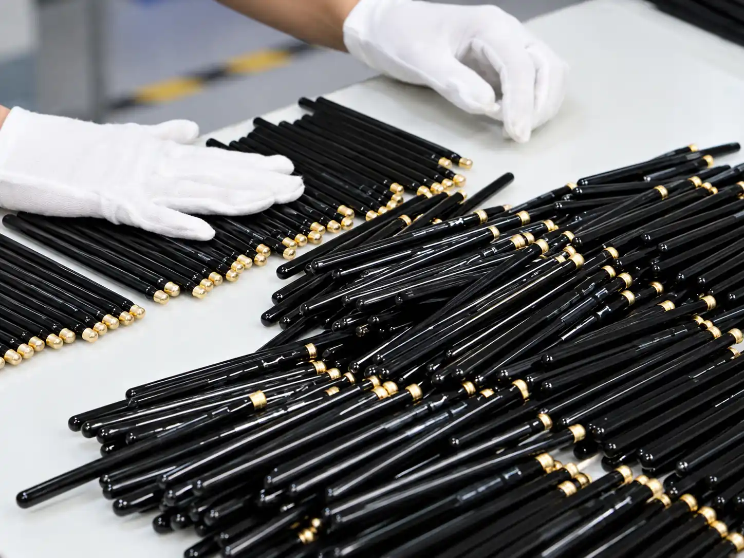 White-gloved hands arranging glossy black makeup brush handles with gold-colored accents