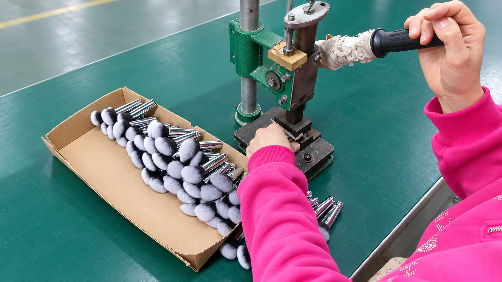 Worker operating a manual press beside a tray of makeup brush heads with silver ferrules