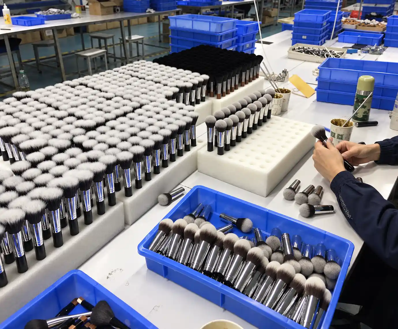 Makeup brush heads arranged in foam holders beside a worker handling brushes at a production table